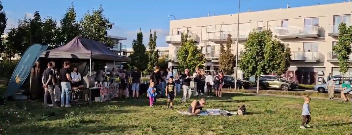 Namiot z poczęstunkiem i grille na pikniku, wokół stoją sąsiedzi, w tle budynki osiedla