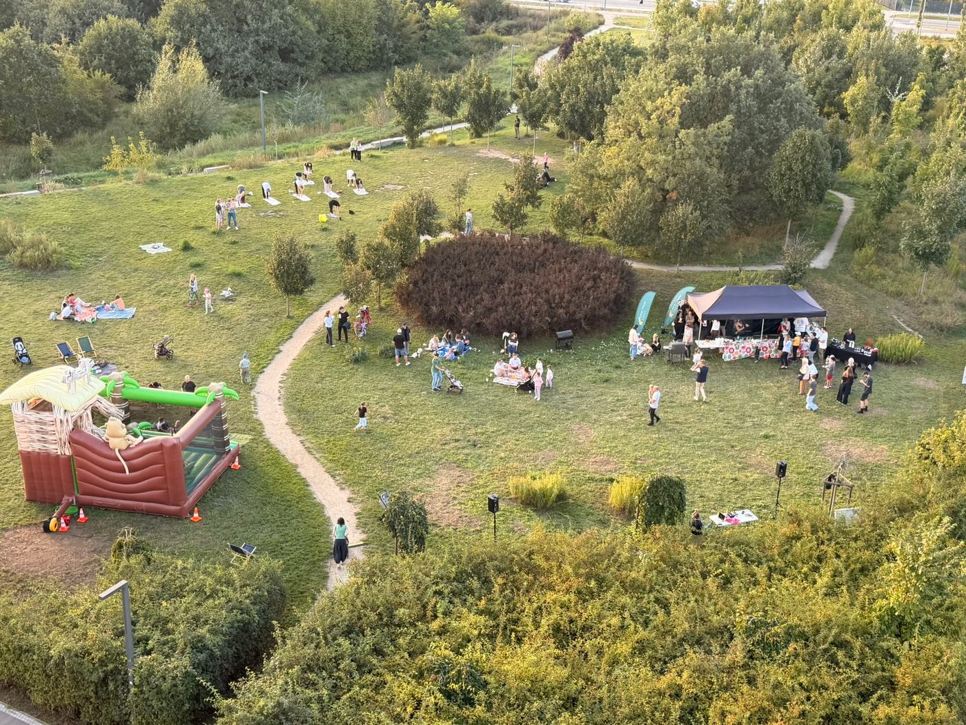 Widok z góry na park podczas pikniku: dmuchany zamek, namiot z grillem, grupa ćwicząca pilates na matach i rozłożone koce